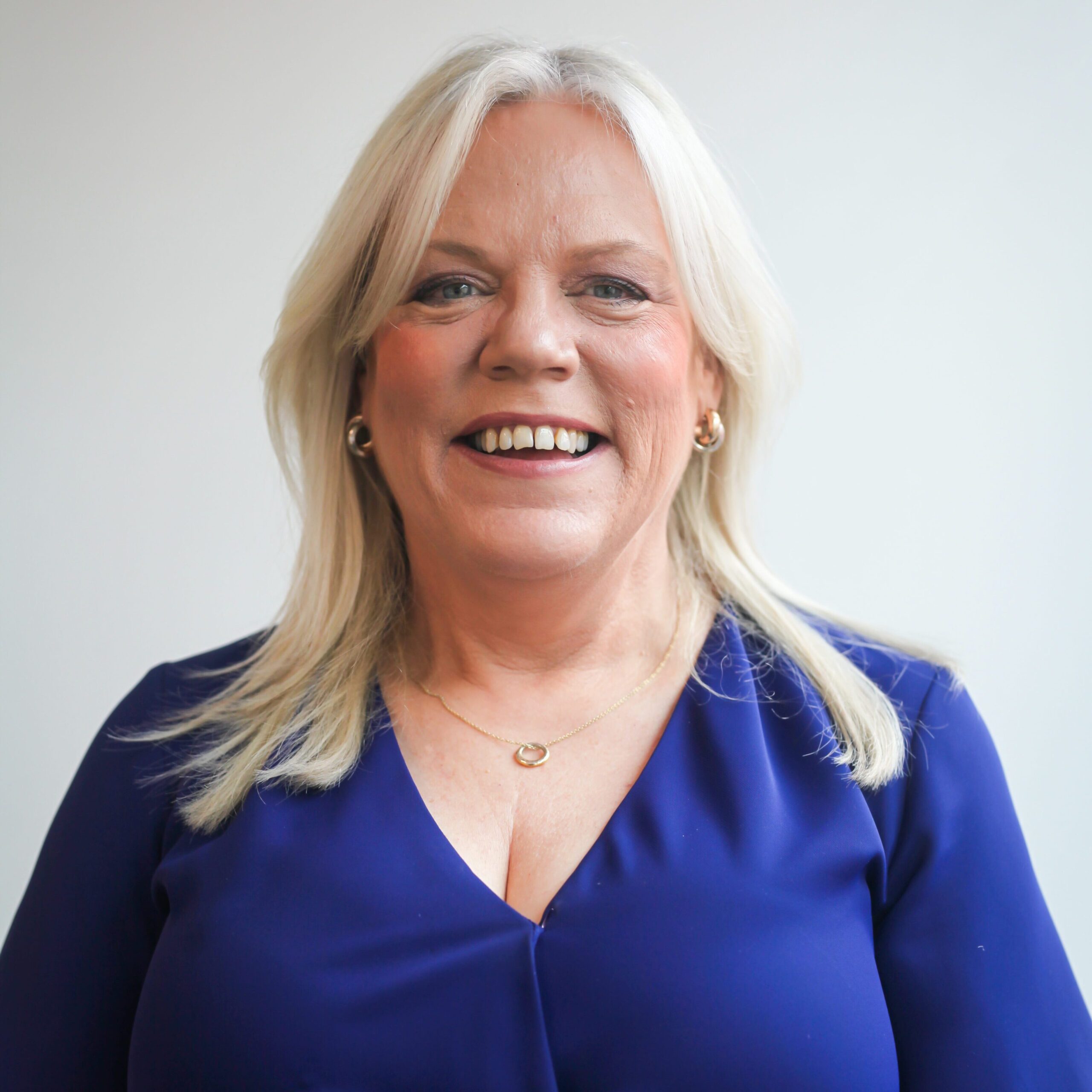 Lorri Weaving the SARSAS CEO. She is smiling and is standing against a white wall backdrop. She wearing a blue top. She is a white women with blonde hair.