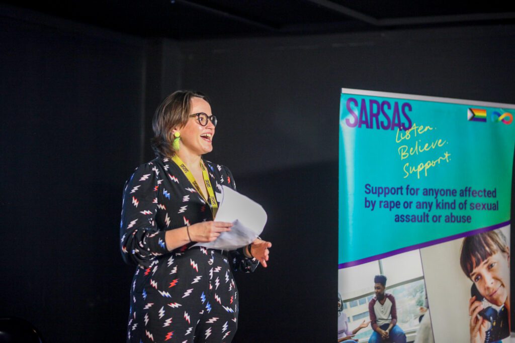 Jenny Benson, Corporate Partnership Lead, standing on a stage next to a SARSAS banner speaking into a microphone