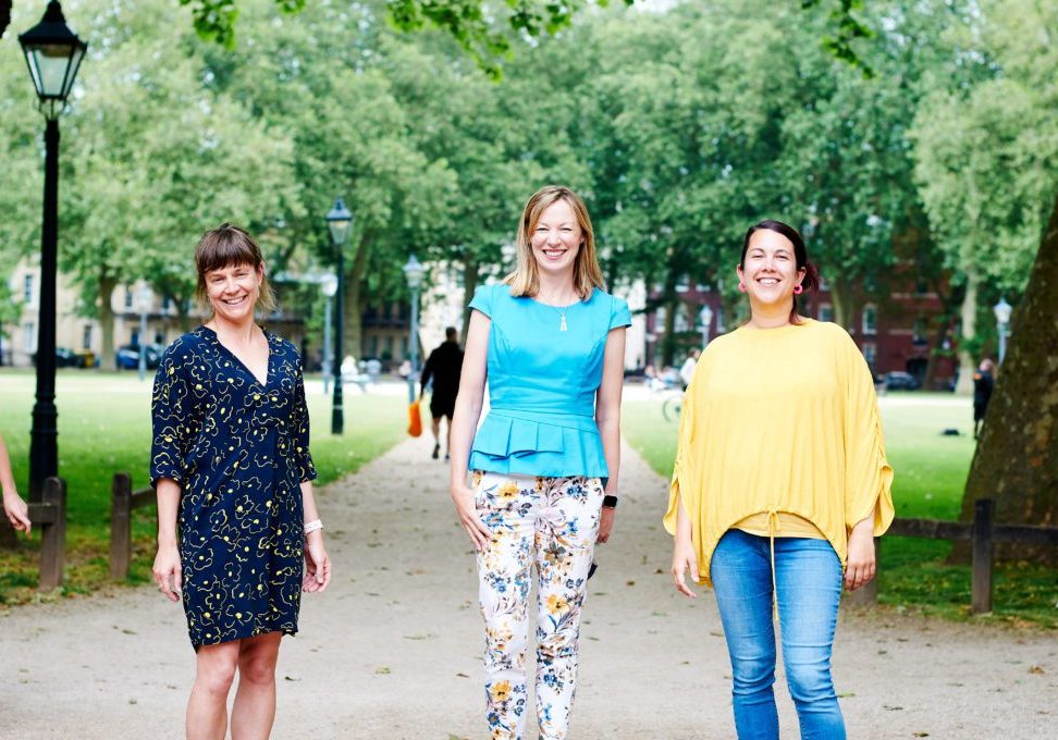 Three women standing in a park. They are wearing colourful clothes and smiling.