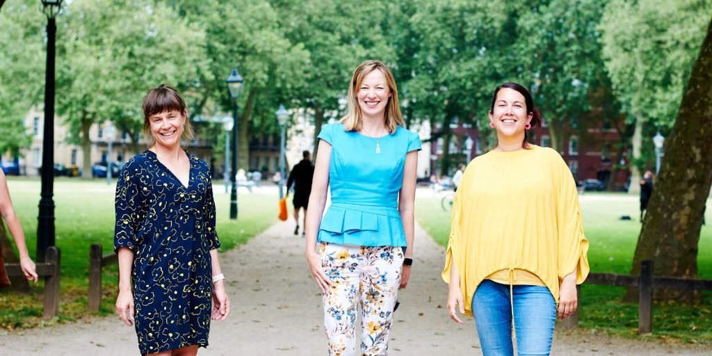 Three women standing in a park. They are wearing colourful clothes and smiling.