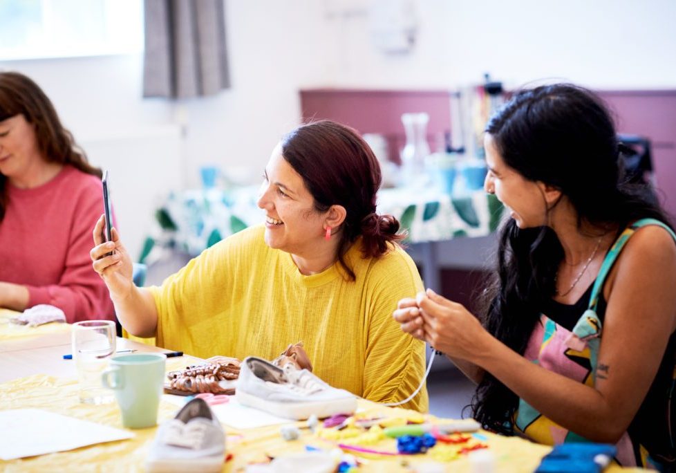 Three women are talking and smiling in a work shop. There are craft supplies in front of them on a table.