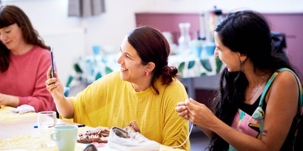 Three women are talking and smiling in a work shop. There are craft supplies in front of them on a table.