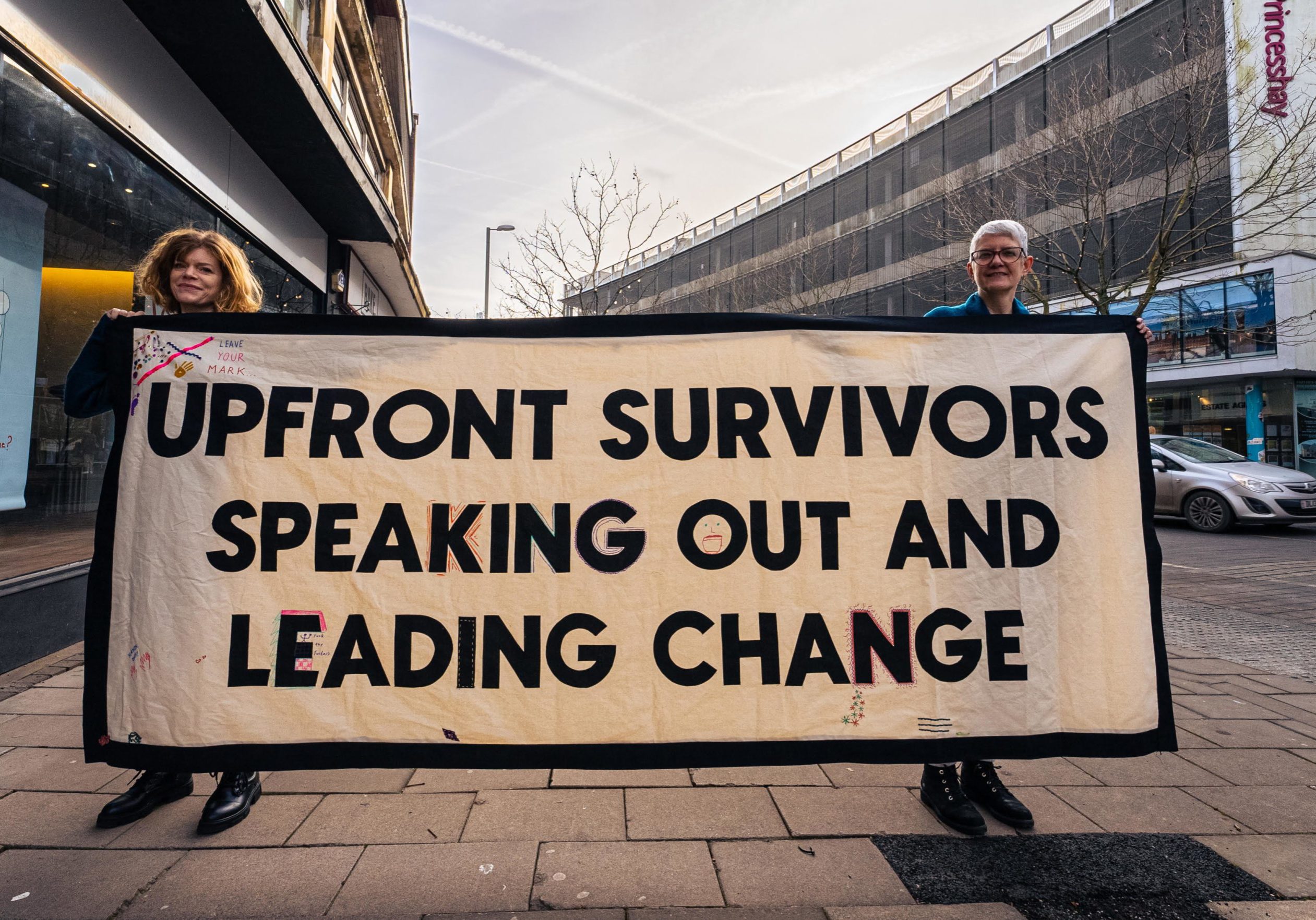 UFS-lead-image.-Photo-by-Fluxx-Films-1 two ladies holding up a sign that says "Upfront survivors speaking out and leading change"