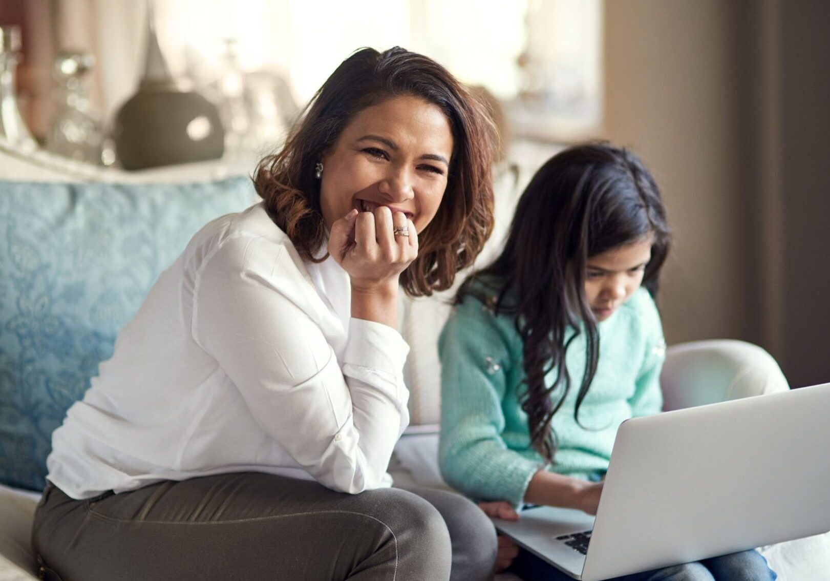 A mother and daughter are sitting on a blue sofa in a living room. The girl is looking at a laptop on her knee and the mother is looking at the camera.
