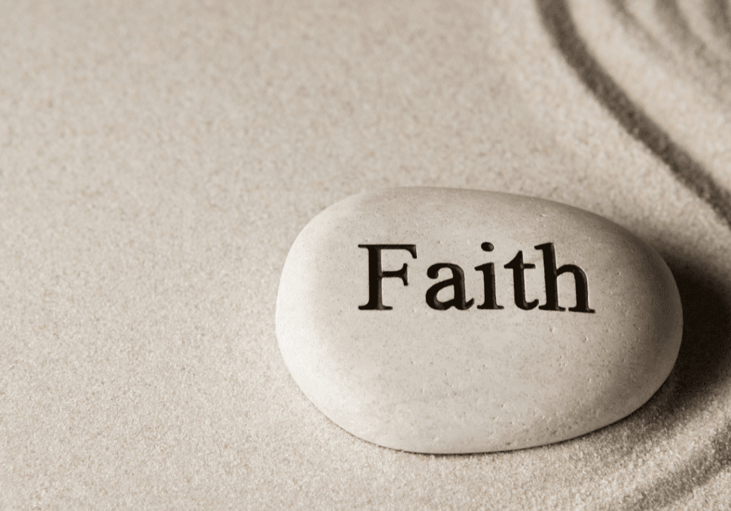 A pebble sits on some beige sand. it has the word faith written on it.