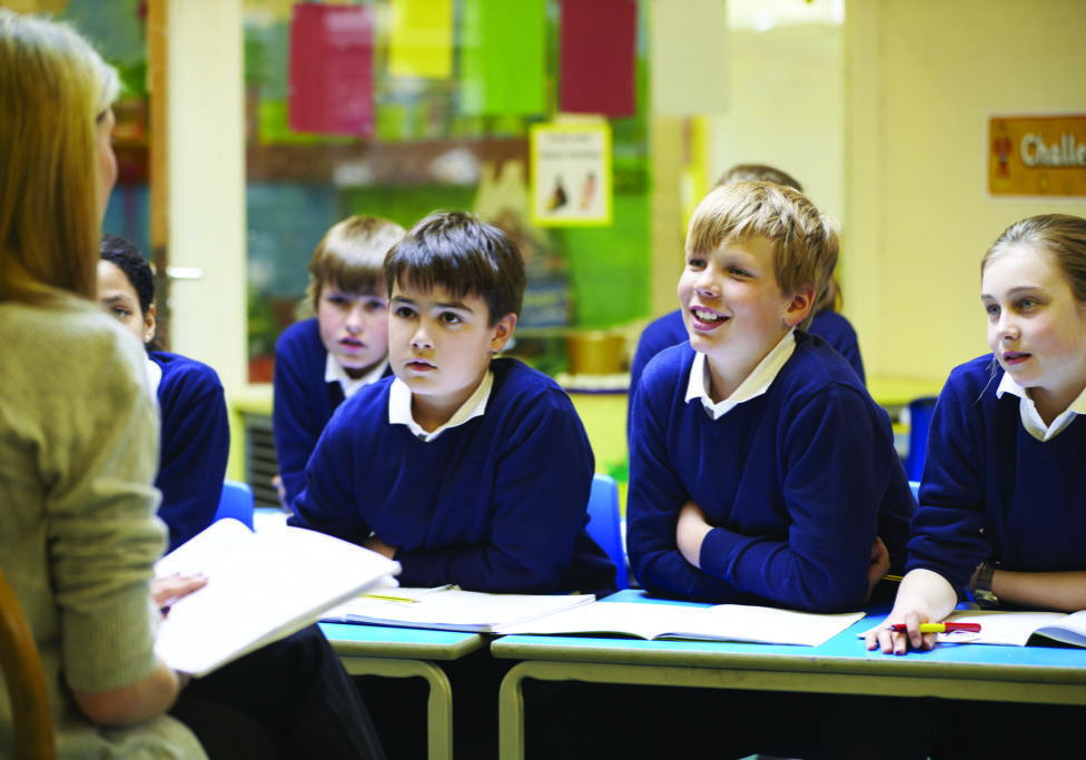 A woman is talking to school children in a classroom. The children are wearing blue uniforms and smiling.