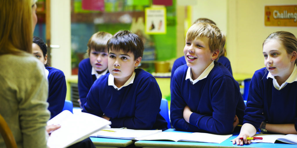 A woman is talking to school children in a classroom. The children are wearing blue uniforms and smiling.